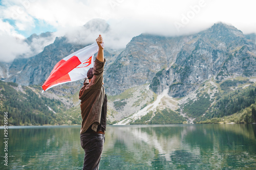man covered in poland flag standing on the edge looking at lake in mountains