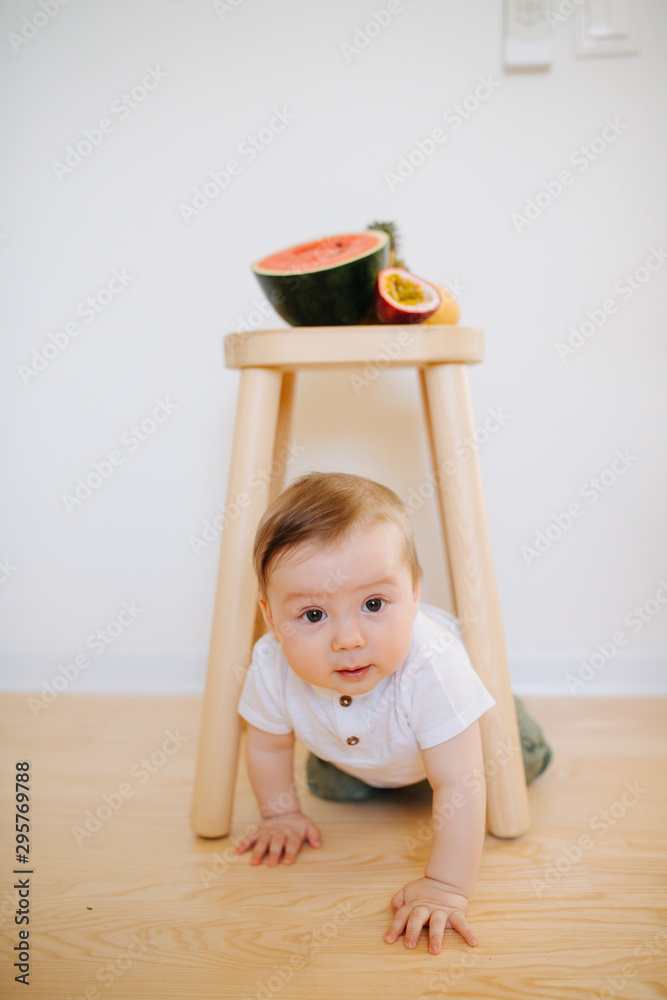 An infant baby is crawling from under the stool with fruits on it An infant baby is crawling from under the stool with fruits on it
