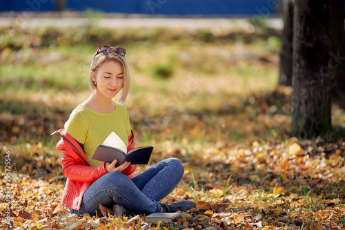 young woman sitting with a book on fallen leaves in the autumn park