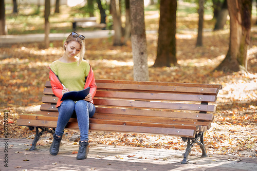 young woman is reading a book while sitting on a bench in a city park. Autumn weather, golden leaves.