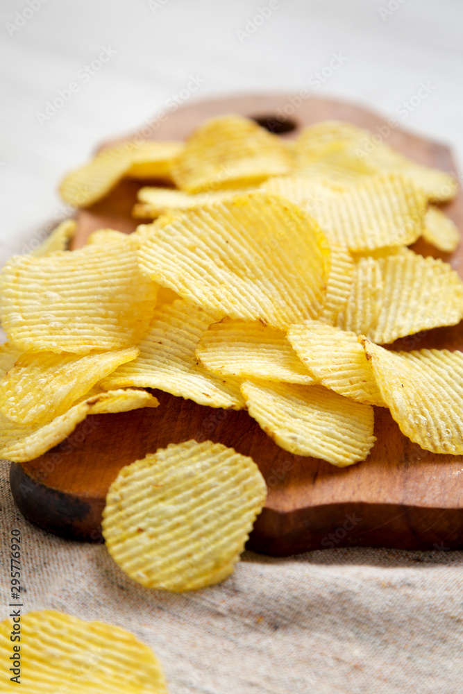 Salted wavy potato chips on a rustic wooden board on a white wooden surface, side view.