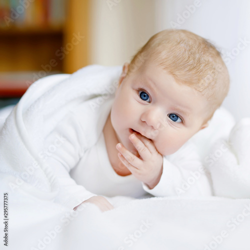 Baby girl with blue eyes wearing white towel or blanket in white sunny bedroom. Newborn child relaxing in bed. Nursery for children. Textile and bedding for kids. Happy cute new born girl or boy.