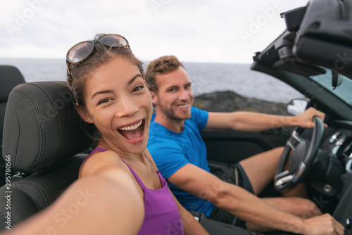 Selfie car travel couple having fun on summer road trip vacation driving to holidays. Asian woman and man excited taking self-portrait photo with mobile phone.