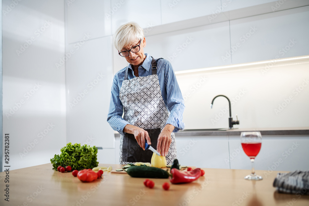 Gorgeous caucasian senior woman in apron shopping pepper and making salad while standing in kitchen. On kitchen counter are vegetables and glass of wine. Gorgeous caucasian senior woman in apron shopping pepper and making salad while standing in kitchen. On kitchen counter are vegetables and glass of wine.