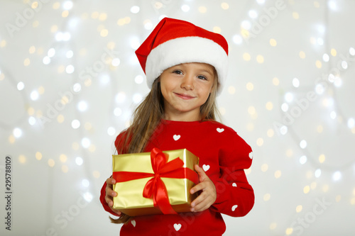 Happy little child in Santa hat with gift box against blurred festive lights. Christmas celebration