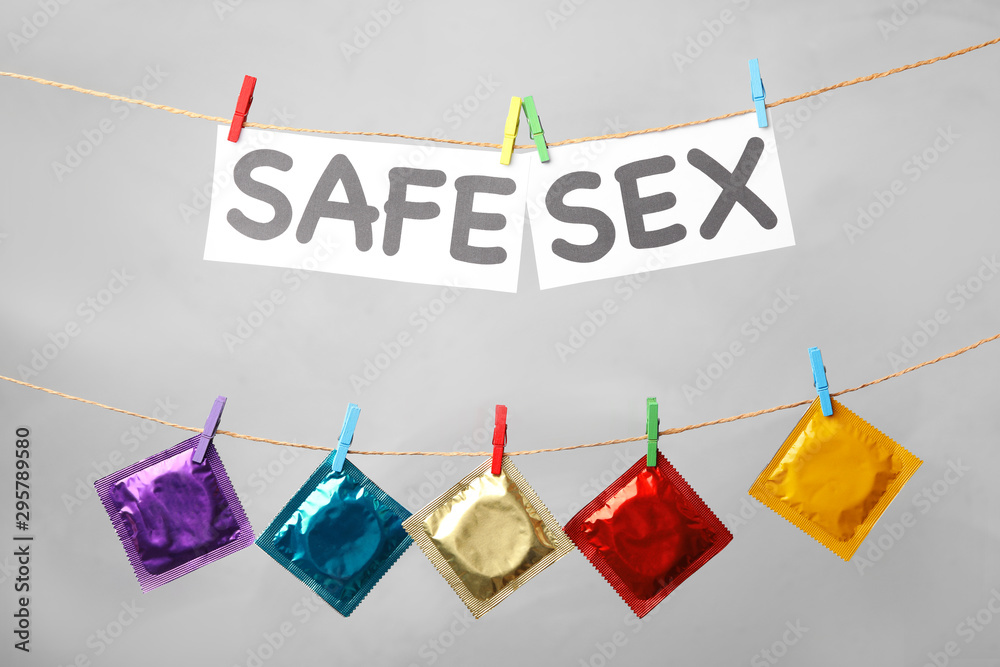 Paper sheets with words SAFE SEX and colorful condoms hanging on clothesline against light grey background - obrazy, fototapety, plakaty 
