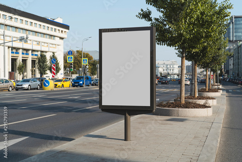 Blank advertizing billboard for commercials in city center. Pedestrian area near busy street with vehicles and cars. Administrative buildings, trees, road signs and multistoried houses in background.