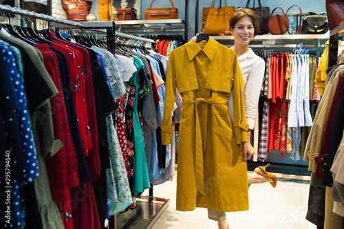 Young woman is demonstrating raincoat of mustard color in clothing shop for women