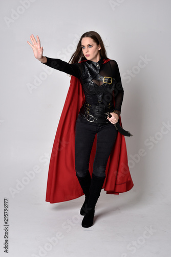 full length portrait of a pretty brunette woman wearing black leather fantasy costume with long red superhero cape. standing pose on a studio background.
