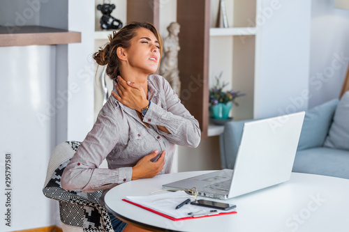 Portrait of young stressed woman sitting at home office desk in front of laptop, touching aching back with pained expression, suffering from backache after working on pc photo