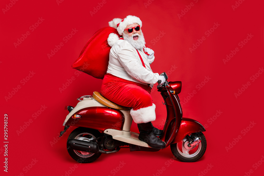 Profile side view portrait of nice bearded cheerful Santa St Nicholas father riding moped hurry up delivering large sack purchase concept isolated on bright vivid shine vibrant red color background Profile side view portrait of nice bearded cheerful Santa St Nicholas father riding moped hurry up delivering large sack purchase concept isolated on bright vivid shine vibrant red color background