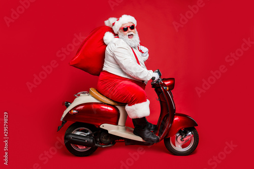 Profile side view portrait of nice bearded cheerful Santa St Nicholas father riding moped hurry up delivering large sack purchase concept isolated on bright vivid shine vibrant red color background