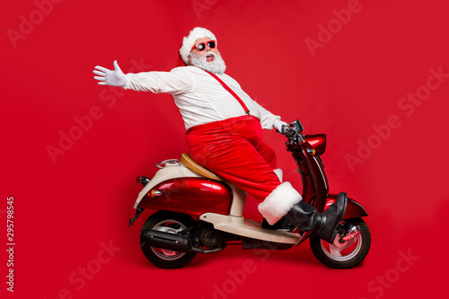 Profile side view portrait of nice bearded glad cheerful cheery funny funky Santa riding moped hurry up having fun holly jolly isolated on bright vivid shine vibrant red color background