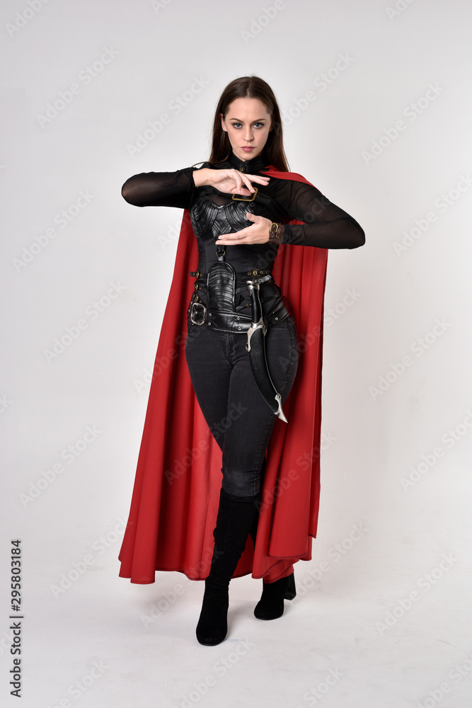 full length portrait of a pretty brunette woman wearing black leather fantasy costume with long red superhero cape. standing pose on a studio background. full length portrait of a pretty brunette woman wearing black leather fantasy costume with long red superhero cape. standing pose on a studio background.