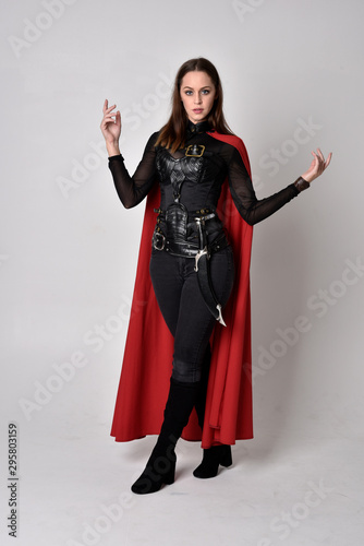 full length portrait of a pretty brunette woman wearing black leather fantasy costume with long red superhero cape. standing pose on a studio background.