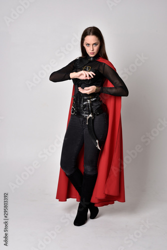 full length portrait of a pretty brunette woman wearing black leather fantasy costume with long red superhero cape. standing pose on a studio background.