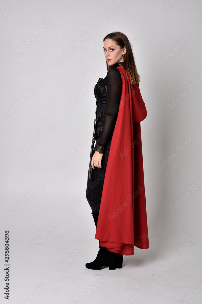full length portrait of a pretty brunette woman wearing black leather fantasy costume with long red superhero cape. standing pose on a studio background. full length portrait of a pretty brunette woman wearing black leather fantasy costume with long red superhero cape. standing pose on a studio background.