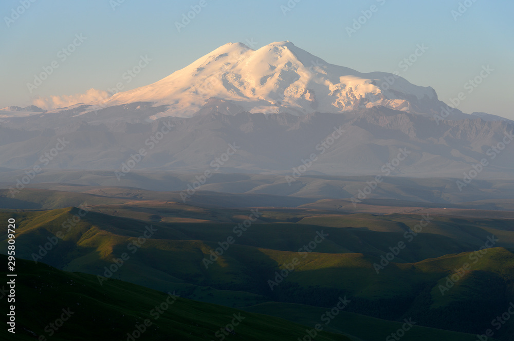 Great nature mountain range. Amazing view of caucasian snow mountain or volcano Elbrus on Bermamyt plateau at sunrise, green fields, blue sky. Elbrus landscape view - highest peak of Russia and Europe