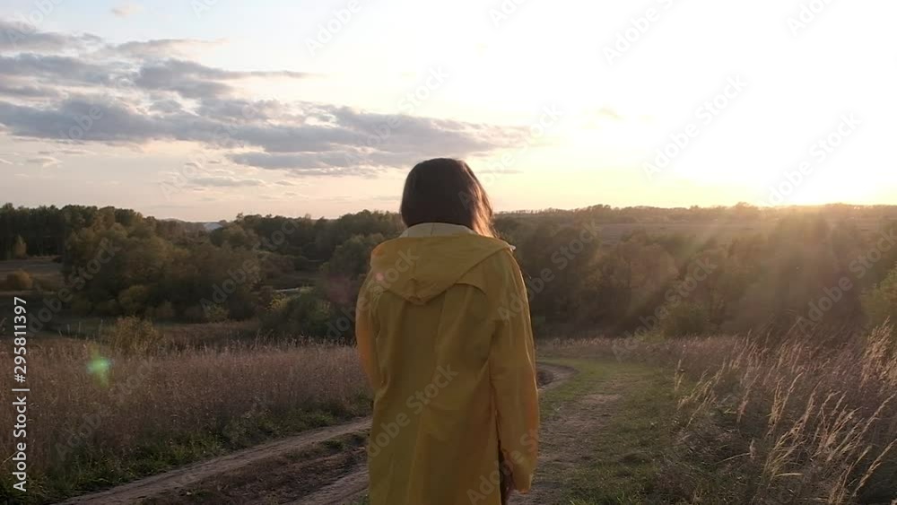 girl in yellow raincoat and rubber boots smiles and calls for walk. woman waves and walks down country road. Sunlight and glare in the camera. Back lighting. Autumn landscape at sunset in September. girl in yellow raincoat and rubber boots smiles and calls for walk. woman waves and walks down country road. Sunlight and glare in the camera. Back lighting. Autumn landscape at sunset in September.