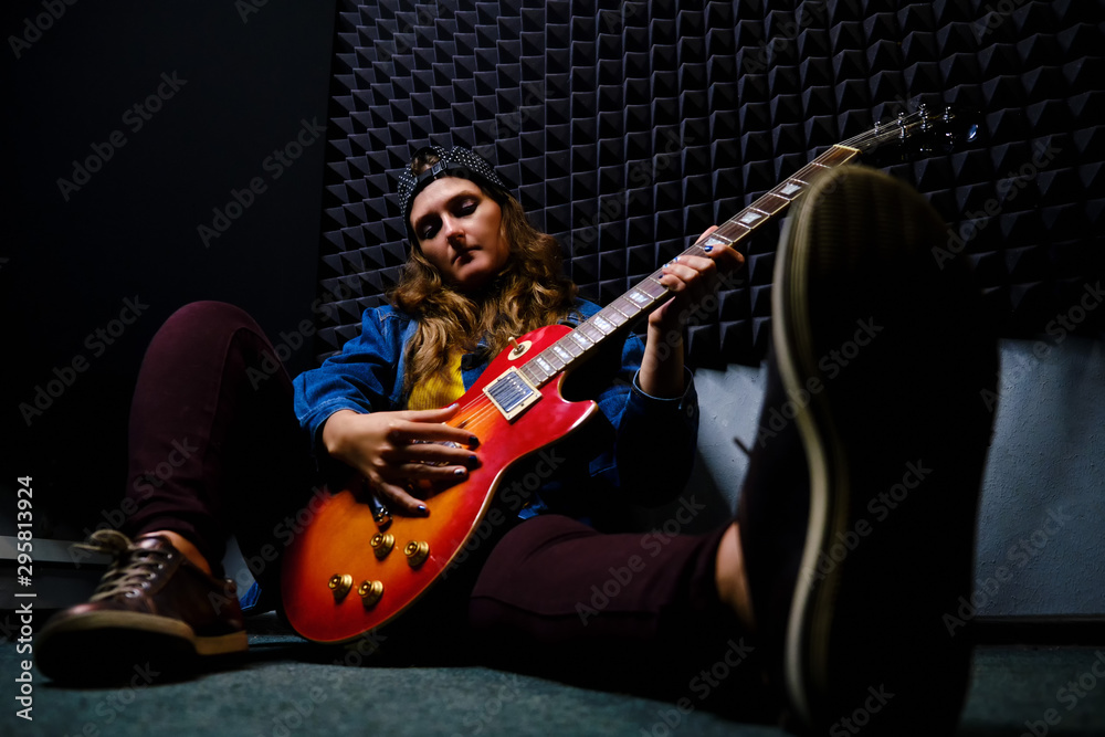 Obraz premium Female guitarist sitting on the floor in a recording Studio. A woman plays music on a red electric guitar. Denim jacket, cap, jeans and sneakers.