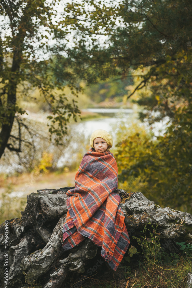 Little girl wrapped in a blanket is sitting on wooden stub near lake in an autumn forest
