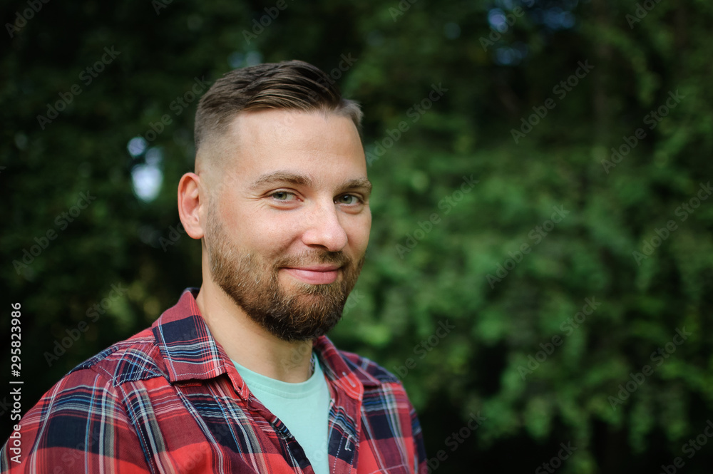 Close up portrait of young bearded man in red shirt looking at the camera with smile in the park on trees background Close up portrait of young bearded man in red shirt looking at the camera with smile in the park on trees background