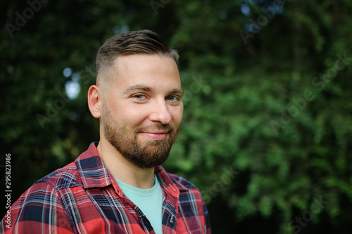 Close up portrait of young bearded man in red shirt looking at the camera with smile in the park on trees background