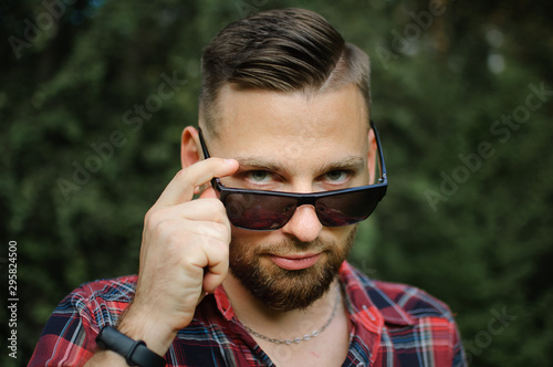 Outdoors portrait of young bearded man with sunglasses looking at the camera in the park on trees background