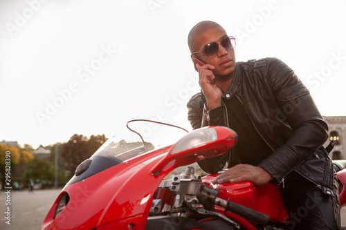 Young African man talking on the phone, resting after riding his sports motorbike, copy space. Handsome male biker using smart phone in the city, sitting on a motorcycle