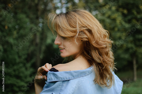 Side view portrait of sexy blonde girl with curly hair wearing blue casual jeans blouse and black lingerie posing in the park on green trees background. Hipster style concept.