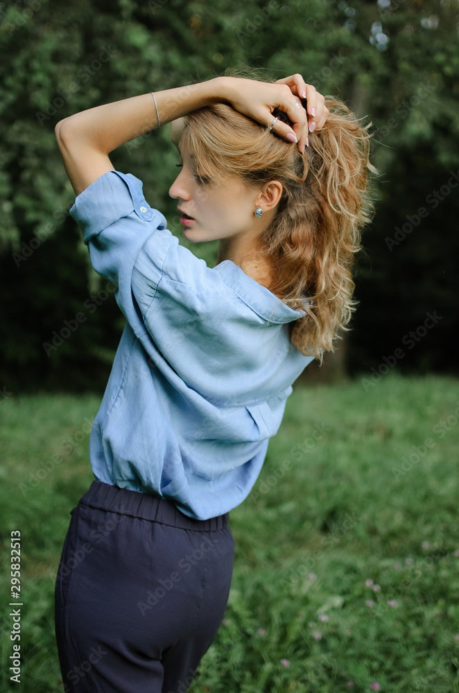 Back view portrait of sexy blonde girl with curly hair wearing blue casual jeans blouse posing in the park on green trees background. Hipster style concept. Back view portrait of sexy blonde girl with curly hair wearing blue casual jeans blouse posing in the park on green trees background. Hipster style concept.