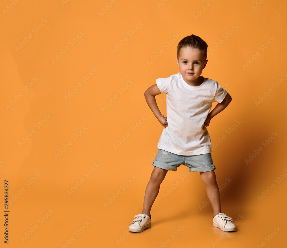 Kid boy in white t-shirt and jeans shorts is standing leaning forward with his hands on his hips at copy space on yellow