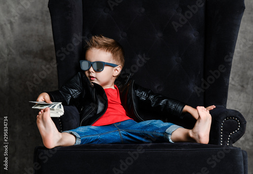 Arrogant kid boy millionaire in leather jacket and red t-shirt is slumped wearily in armchair holding a bundle of cash