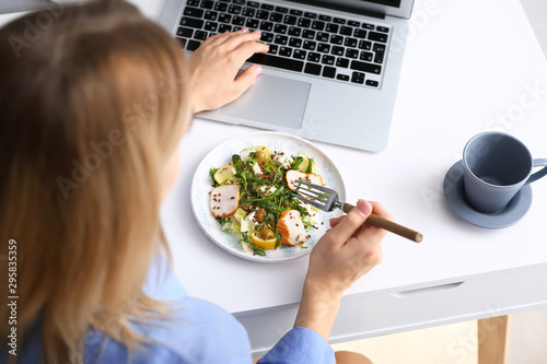 Woman eating tasty salad while working on laptop at home