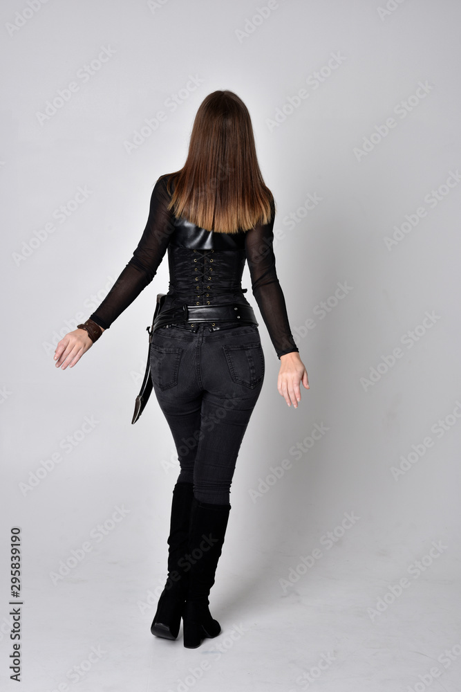 full length portrait of a pretty brunette woman wearing black leather fantasy costume with a dagger. standing pose, facing away from camera on a studio background.