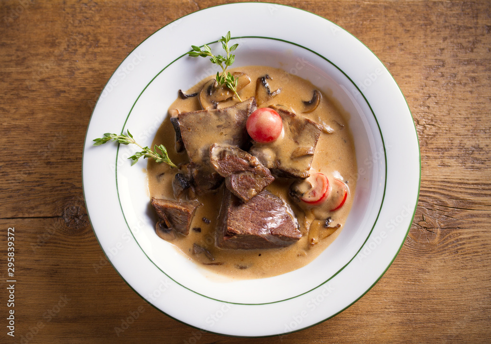 Braised beef cheeks in cream sauce with mushrooms and tomatoes in white bowl on wooden table. Overhead