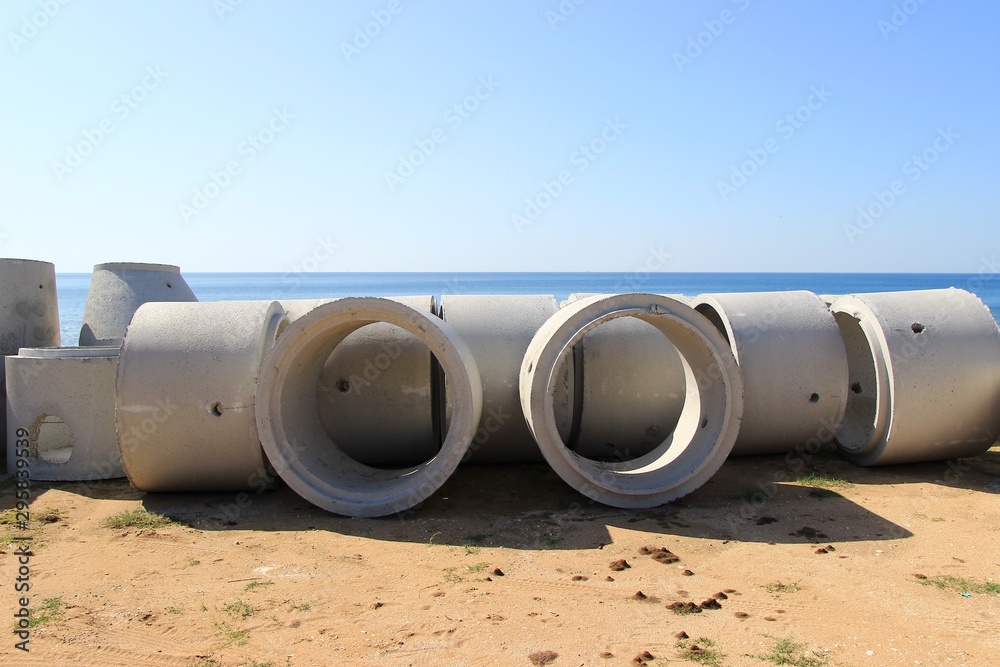 Large new concrete circular pipes lying down on a sandy beach for the construction of sewerage system background view of clear blue sea and sky on a sunny day Large new concrete circular pipes lying down on a sandy beach for the construction of sewerage system background view of clear blue sea and sky on a sunny day