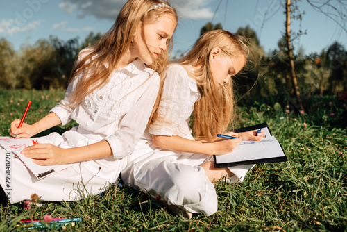 two little girls with pencil draws in notebook while sitting on the grass in the park, child writes in notebook