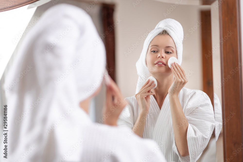 A cute girl in a white waffle bathrobe with a towel on her head poses while standing in front of a large mirror and removing makeup residue from her face with cotton pads. A cute girl in a white waffle bathrobe with a towel on her head poses while standing in front of a large mirror and removing makeup residue from her face with cotton pads.