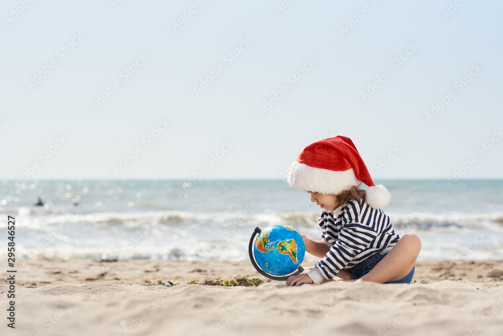 Obraz premium Happy little child girl in Santa hat sits on sand with globe on tropical beach. Kid having fun on summer vacation on Christmas and New year. Holiday Travel and adventure concept