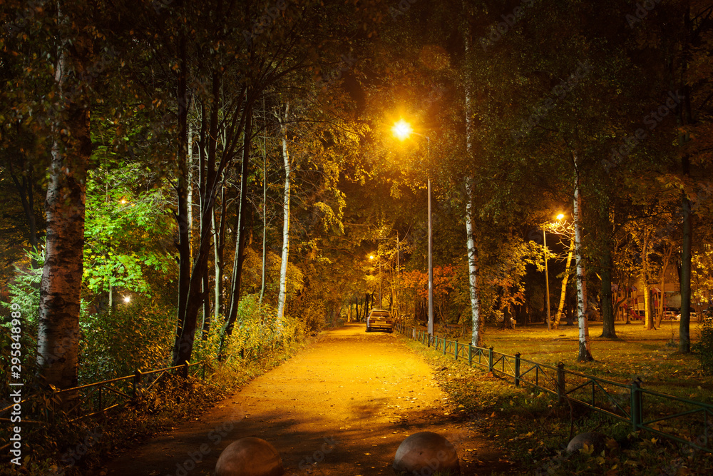 Deserted, nightly path lit by a lantern in an autumnal city courtyard among the trees of a small square. A lone car parked in the distance. Night shooting.
