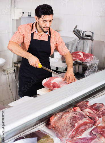 Adult butcher is cutting meat for seller to clients indoors.