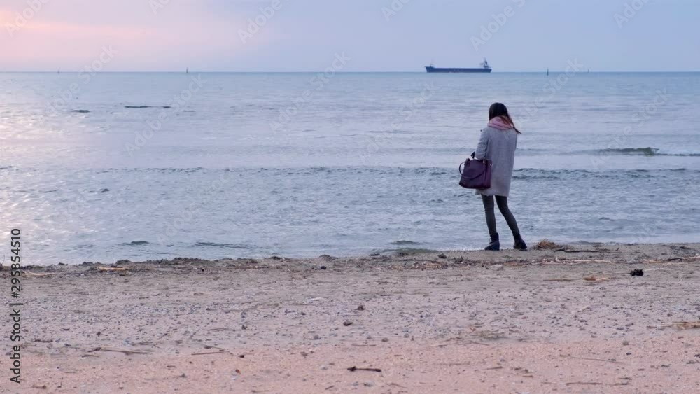 Young woman admires sea view on sunset at sea beach in winter, back view. Girl in grey coat standing at seashore and looking at distnce. Traveler, travel, journey, tourism, trip on vacation. Young woman admires sea view on sunset at sea beach in winter, back view. Girl in grey coat standing at seashore and looking at distnce. Traveler, travel, journey, tourism, trip on vacation.