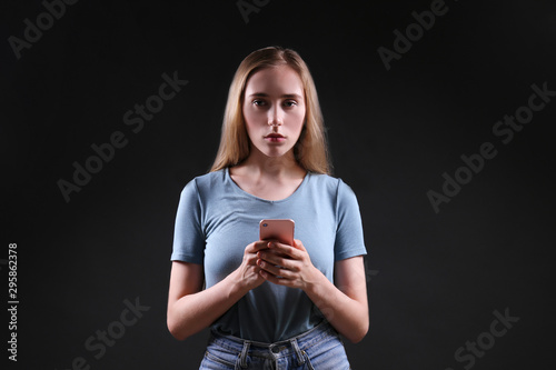 Portrait of beautiful young woman with depressed facial expression standing in the dark holding her phone. Cyber bullying victim concept. Sad female staring at smartphone screen. Background copy space