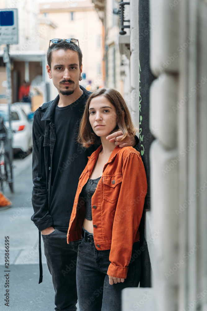 Vertical photo of a couple in the street of a city embraced by the back and looking at camera