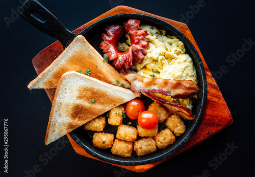 Top view image of western brunch menu on pan and wooden plate. American style breakfast served with toast, hash brown, sausages,bacon, scrambled eggs, and tomatoes on a black background.