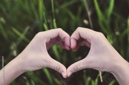The person hand shows the heart shape symbol on the grass background