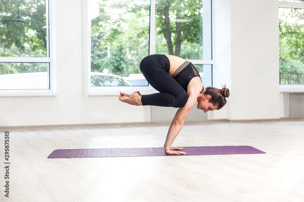 Full lenght side view portrait of young adult sporty attractive woman in black pants and top is practicing yoga, standing in crane, bakasana pose, working out, indoor, window background