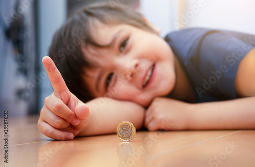 Blurry image of Kid face laying dow on wooden floor showing money coin, Selective focus of happy child pointing finger up and playing with one pound coin. Positive children concept