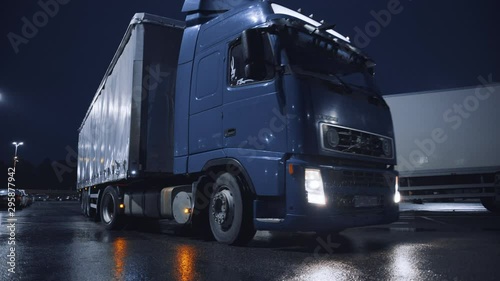 Blue Semi-Truck with Cargo Trailer Drives Off From Overnight Parking Space where Other Trucks are Standing. Long Haul Truck Leaves Parking Lot, Transporting Cargo / Goods Across Continent. Rainy Night